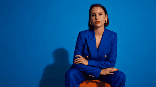 Mujer con traje azul sentada sobre un banco naranja frente a un fondo azul, con brazos cruzados suavemente y mirada directa a cámara.