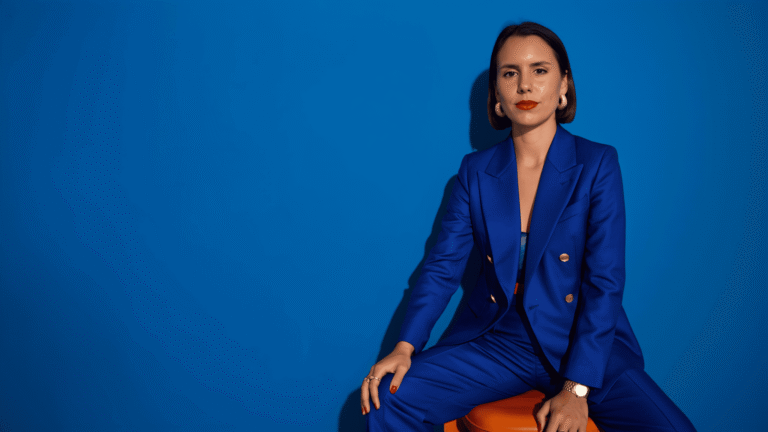 Mujer sentada frente a un fondo azul intenso, con traje azul y labios rojos, mirando a cámara con expresión seria.