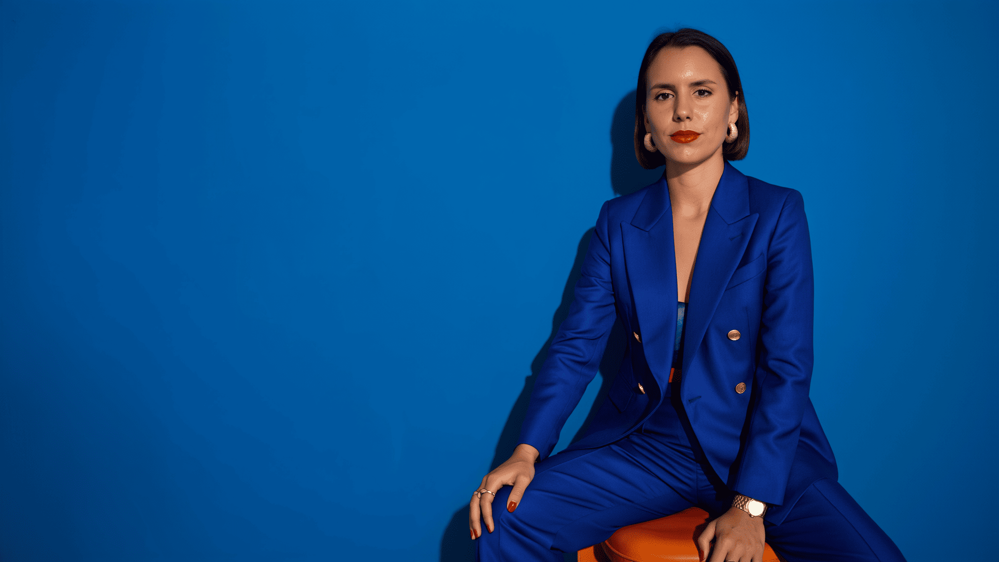 Mujer sentada frente a un fondo azul intenso, con traje azul y labios rojos, mirando a cámara con expresión seria.